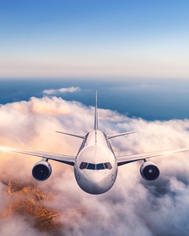 Airplane is flying over clouds at sunset in summer. Landscape with passenger  airplane, low clouds, sea, purple sky at dusk. Front view of the aircraft. Business travel. Commercial plane. Aerial view